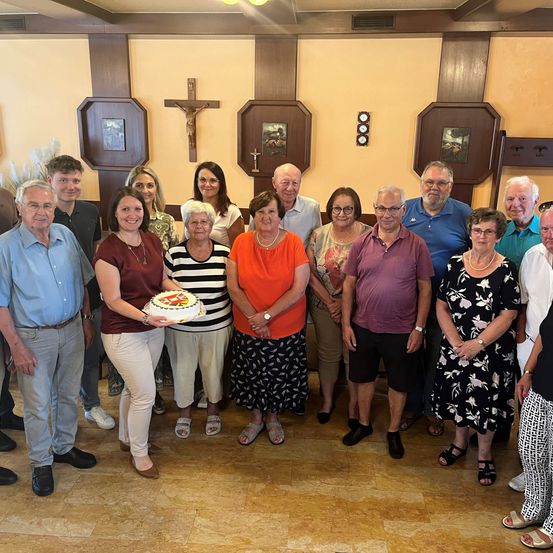 Eine Gruppe von Menschen steht für ein Foto in einem Raum, einige mit Brille und lächelnd. Eine Frau hält einen Kuchen, und ein Kreuz ist an der Wand.