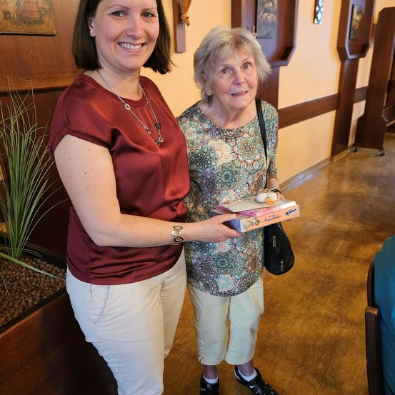 Zwei Frauen stehen in einem Flur und lächeln. Eine hält eine Schachtel Pralinen, die andere eine Handtasche. Im Hintergrund sind eine Pflanze und Bilderrahmen zu sehen.