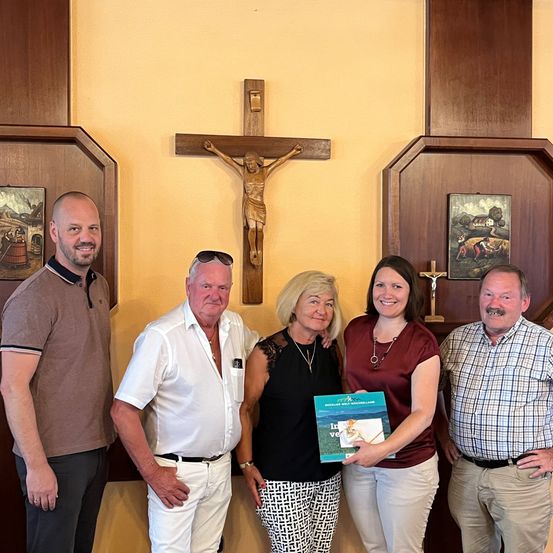Fünf Personen posieren für ein Foto in einer Kirche. Eine Frau hält ein Buch. Hinter ihnen schmücken ein Kreuz und zwei Gemälde die Wand.