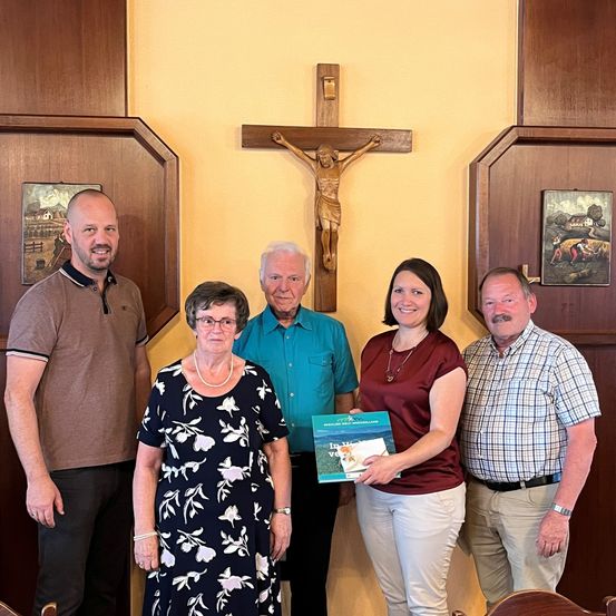 Fünf Personen stehen in einem gelb gestrichenen Raum und posieren für ein Foto. Eine Frau hält ein Buch. Dahinter ist ein Kruzifix an der Wand angebracht.