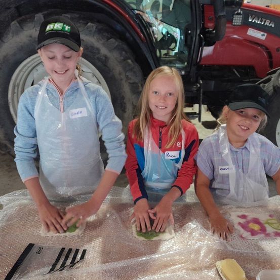 Drei Mädchen arbeiten an einem mit Plastik bedeckten Tisch auf Teig. Ein Mädchen hat einen Namensschild, auf dem Linda steht. Ein Traktor ist im Hintergrund.