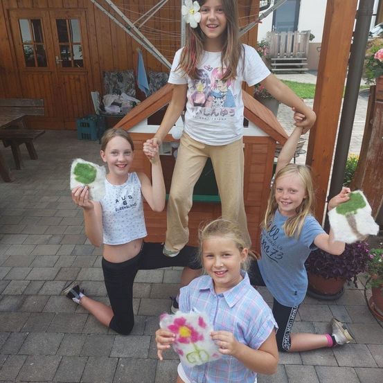 Drei junge Mädchen posieren mit selbstgemachten Kunstwerken im Freien. Ein Mädchen steht auf den Schultern einer anderen. Sie halten selbstgemachte Blumen, mit einem kleinen Haus im Hintergrund.