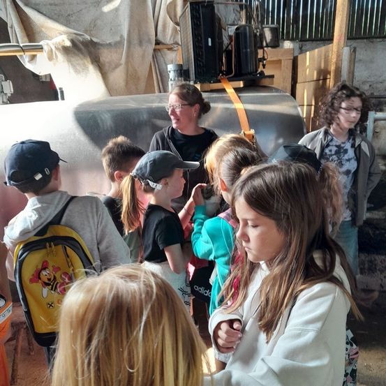 Mehrere Kinder sind in einer Scheune um eine Frau versammelt. Sie betrachten einen großen Metalltank mit Maschinen obenauf. Die Frau lächelt und spricht mit ihnen.