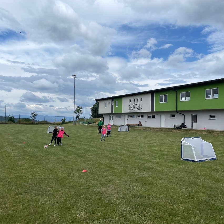 Eine Fußballtrainingseinheit findet auf einem Rasenfeld statt, mit einem grünen Gebäude im Hintergrund. Mehrere junge Spieler, die pinke und schwarze Trikots tragen, treten einen Fußball.