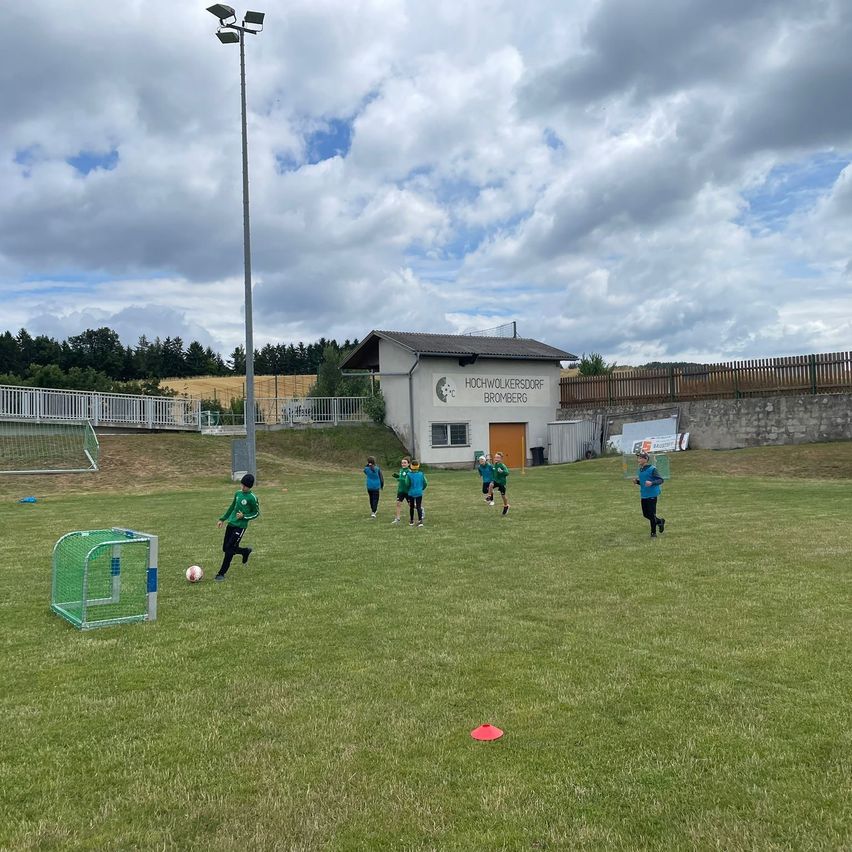 Eine Gruppe von Kindern spielt Fußball auf einem Feld. Im Hintergrund befindet sich ein Gebäude mit einem Schild, das Hochwolkersdorf Bromberg besagt.