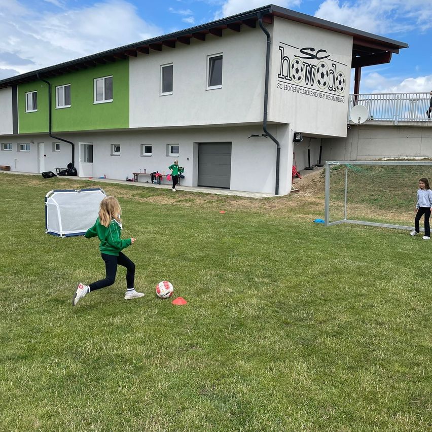 Ein junges Mädchen übt Fußballtritte auf einem grünen Feld vor einem Gebäude mit der Aufschrift 'SC Hochwolkersdorf Bromberg'. Ein anderes Mädchen beobachtet sie.