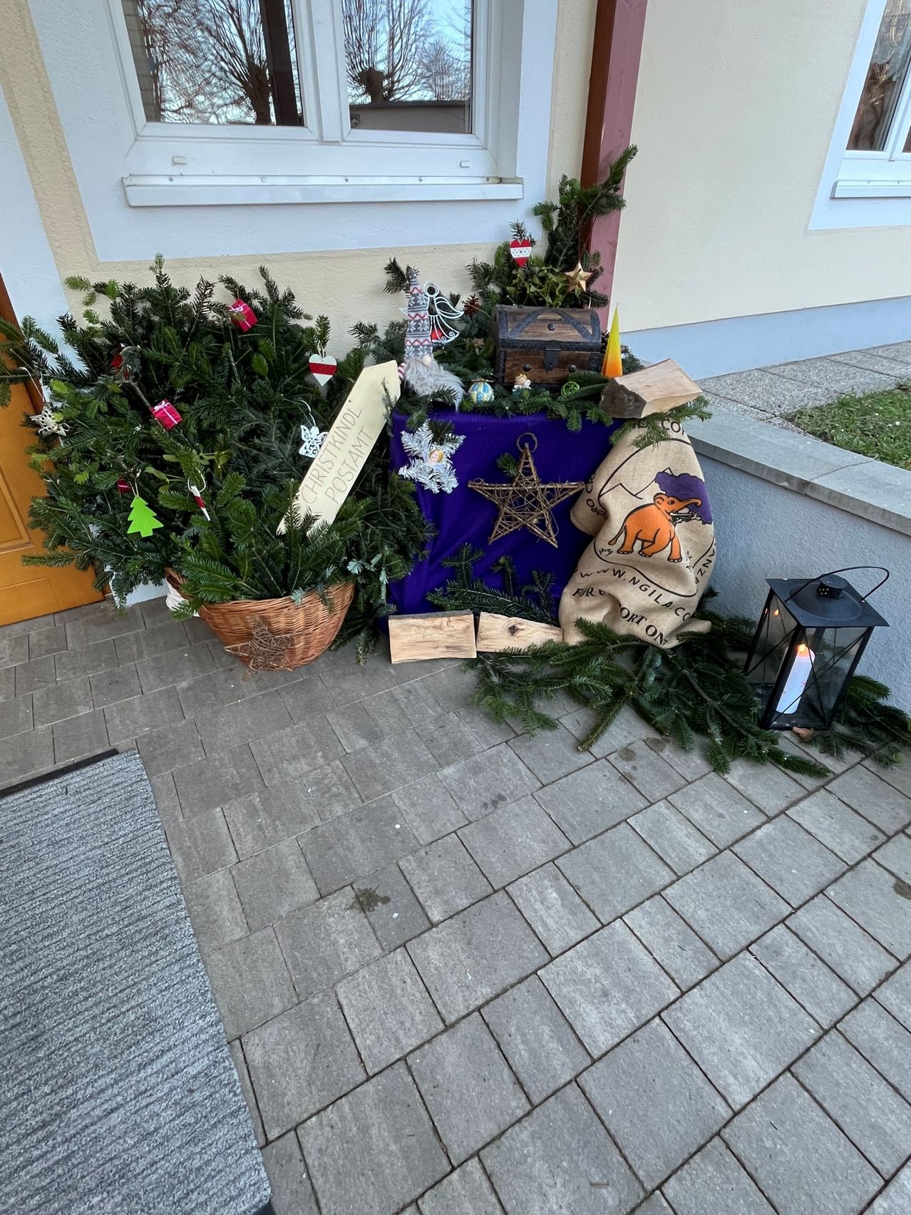 Eine Weihnachtsbaum-Arrangement steht auf einem Kiesweg. Es besteht aus einem Korb mit Tannenzweigen, einem Holzkasten und einem Stern auf einem lila Tuch. Ein Laterne ist in der Nähe.