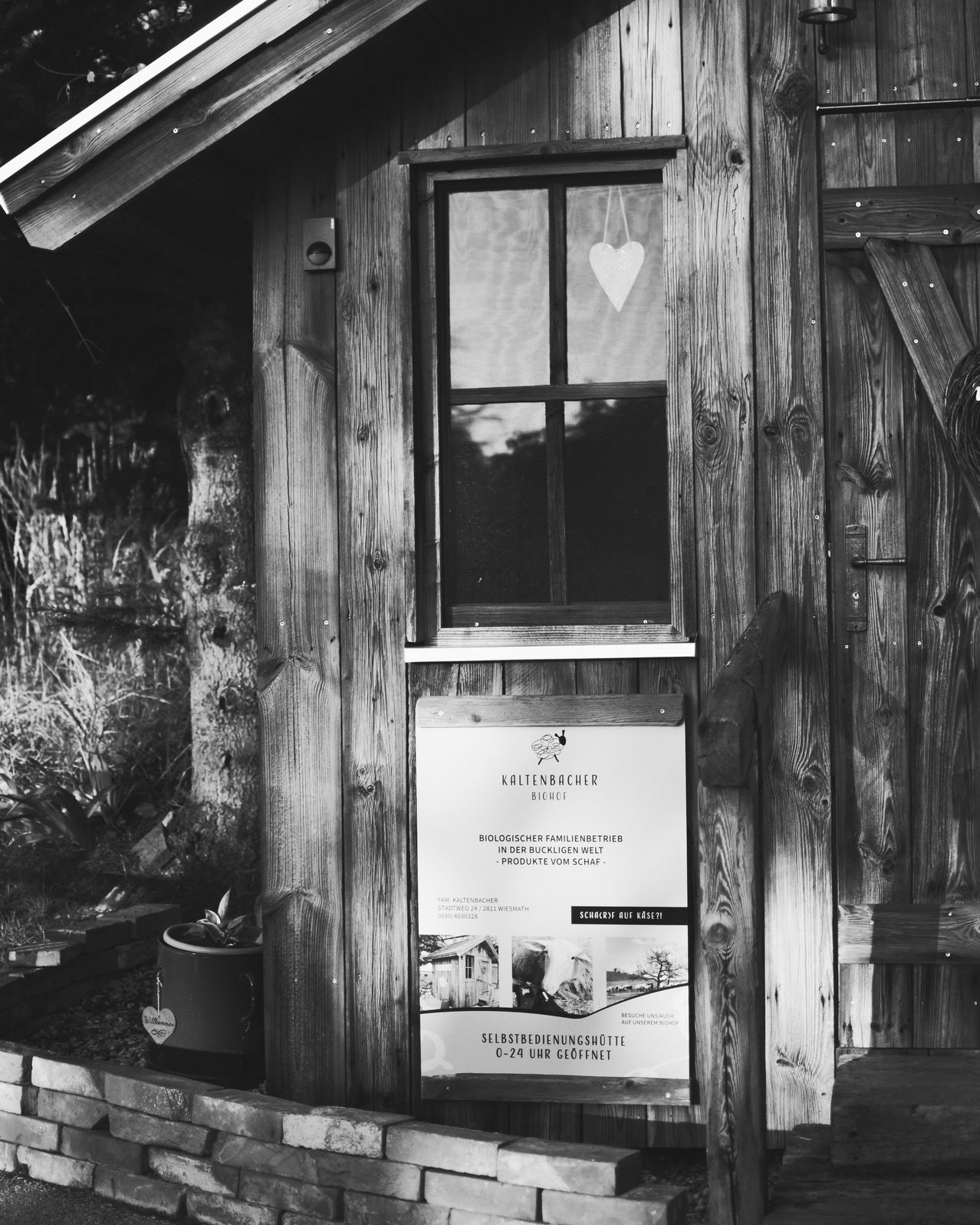 Schwarz-Weiß-Bild einer Holzhütte. Sie hat ein Fenster, eine Tür und ein Schild mit der Aufschrift Kaltenbacher. Ein Herz hängt im Fenster. Ein Topf und ein Herz liegen auf dem Boden.