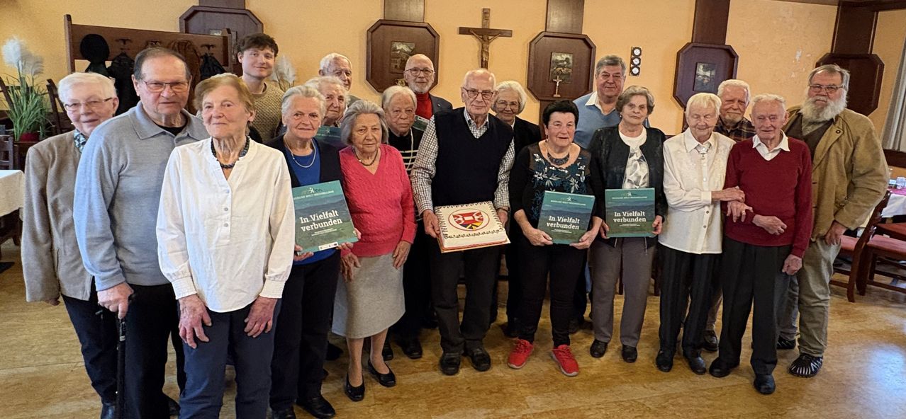 A group of elderly individuals stand together in a room, holding books titled 'In Vielfalt Verbunden'. They pose for a photo, smiling and looking at the camera.