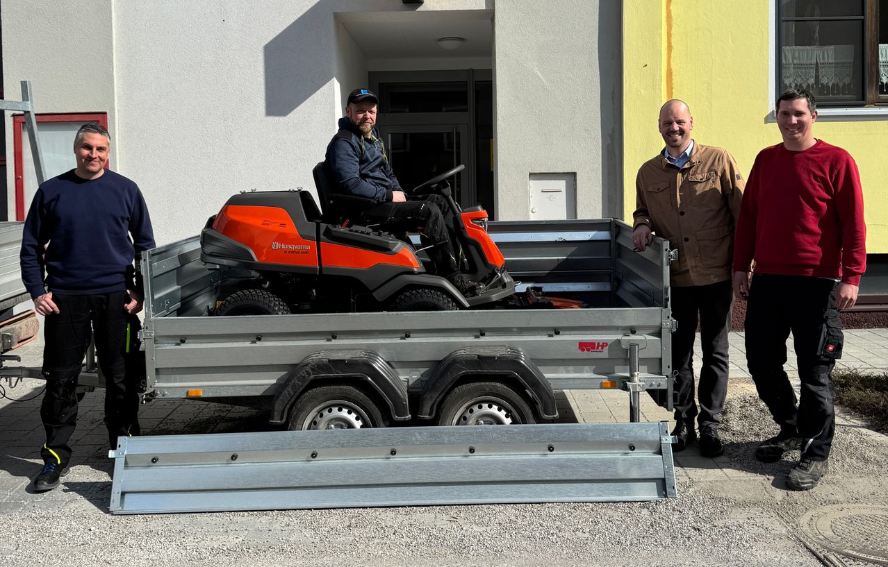 Zwei Männer stehen neben einem Anhänger mit einem roten Rasenmäher darauf. Ein Mann sitzt auf dem Rasenmäher. Sie lächeln beide.