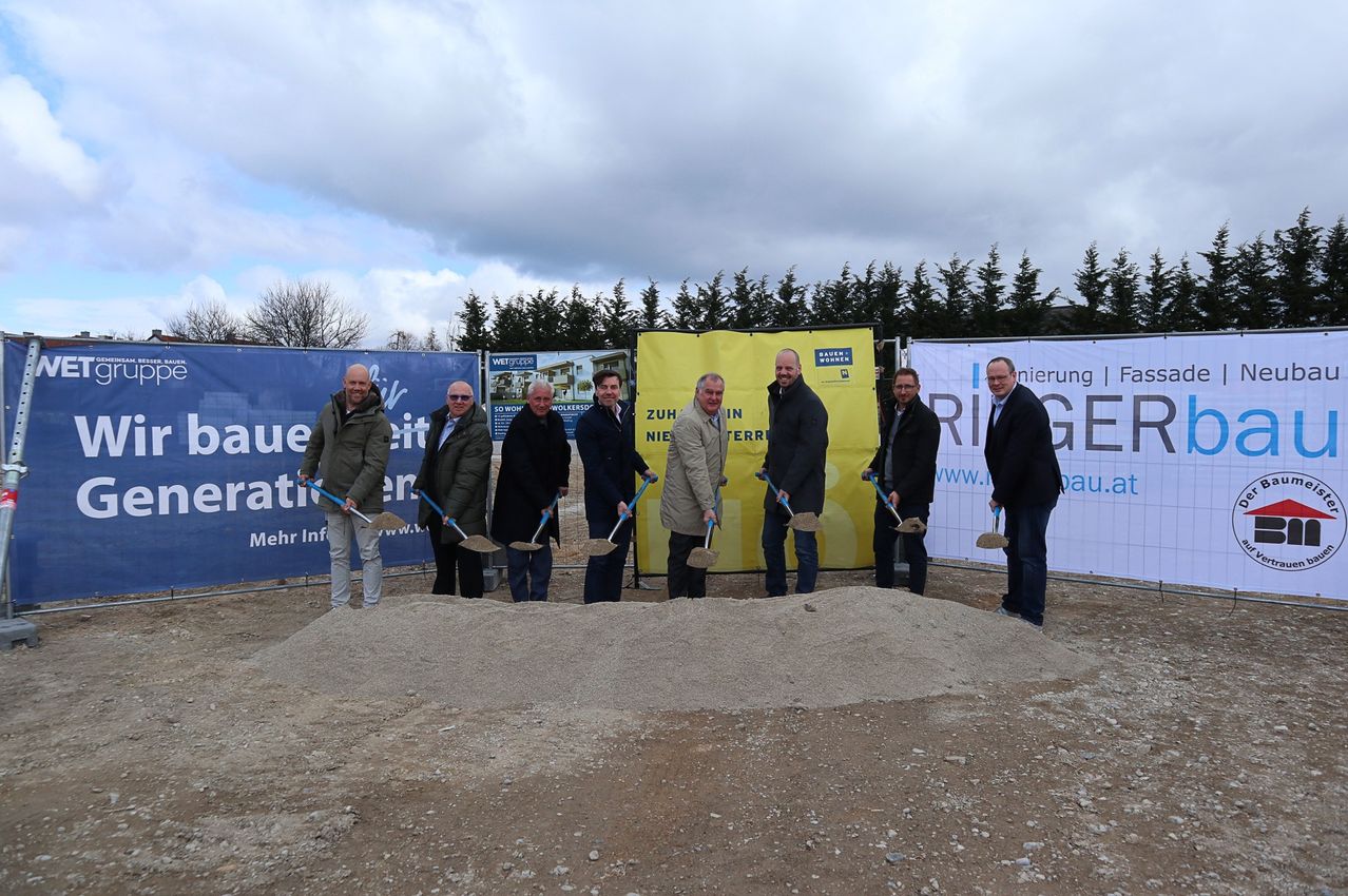 Sieben Männer stehen um einen Haufen Erde herum und halten Schaufeln in der Hand. Sie sind in Jacken und Hosen gekleidet und stehen vor einer Baustelle. Hinter ihnen befinden sich Banner, von denen eines das Wort 'Ries' darauf hat.