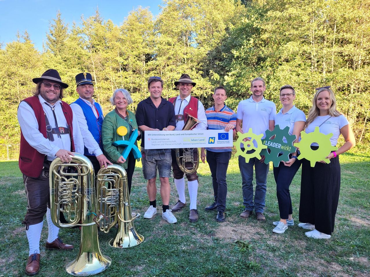 A group of people in traditional Bavarian attire are posing for a photo with a large check and musical instruments. They are standing in a grassy area with trees in the background.