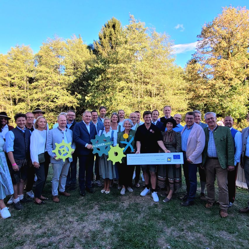 A group of people stands in a park with a large check, smiling and holding green objects. Trees and a blue sky are in the background.