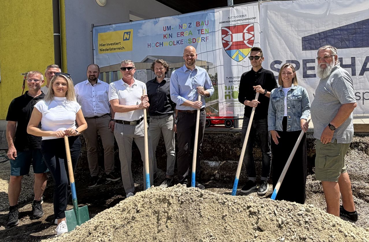 Mehrere Personen stehen mit Schaufeln an einer Baustelle, lächeln und posieren für ein Foto. Ein großes Banner hinter ihnen trägt die Aufschrift 'UM N DZ BAU KIN ERGA TEN HOCHWOLKERSD'.
