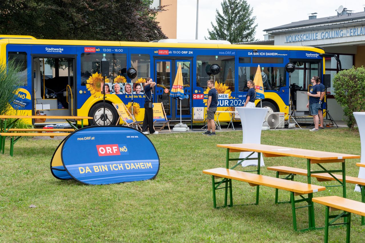 Ein blauer Bus mit Radiosendermarke steht auf einem Rasen. Menschen stehen in der Nähe des Busses mit offener Tür und zwei Frauen sind im Inneren.