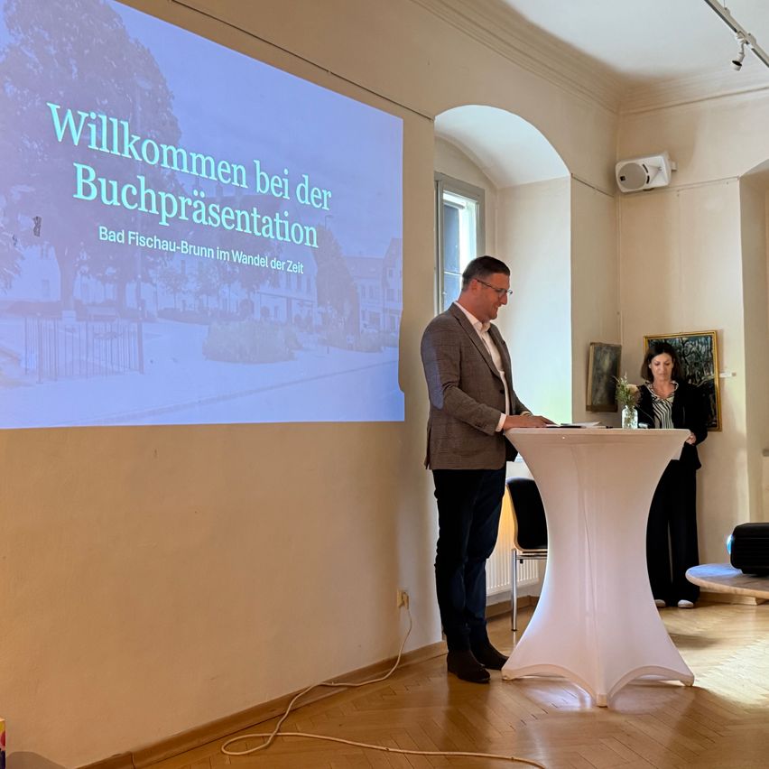 Ein Mann hält eine Präsentation an einem Podium vor einer Projektionsleinwand, auf der Willkommen zur Buchpräsentation steht.
