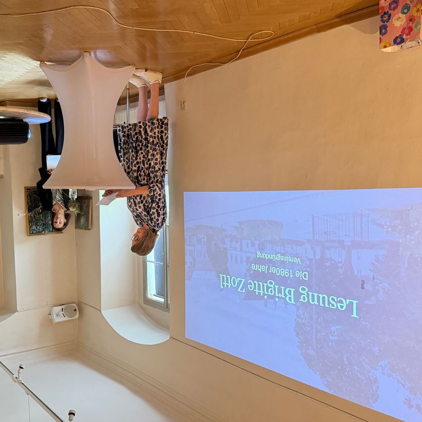 Eine Frau hängt kopfüber mit den Füßen an der Decke. Eine Projektionsleinwand zeigt 'Zukünftiges Stadtdesign'. Der Raum hat eine weiße Decke und Wände.