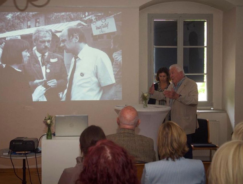 Eine Präsentation mit projiziertem Bild zeigt zwei Männer in Anzügen. Ein älterer Mann spricht an einem Tisch mit einem Blumenvase. Eine Frau steht in der Nähe. Leute sitzen und schauen zu.