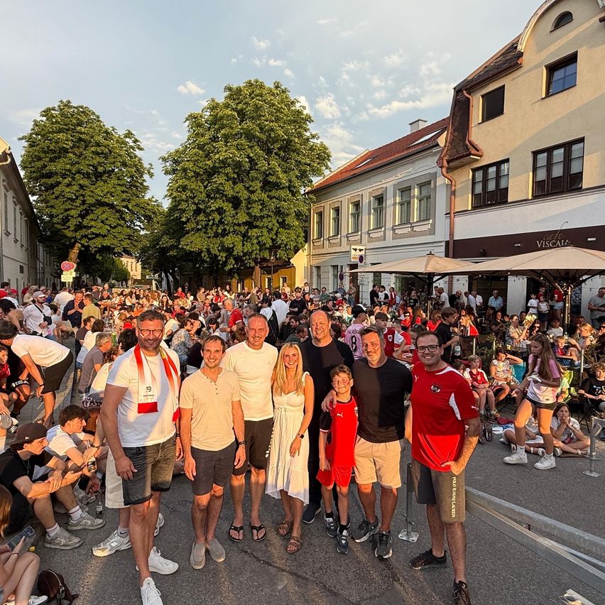 Eine Gruppe von Menschen, einschließlich Erwachsener und Kinder, steht gemeinsam in einer Outdoor-Umgebung, möglicherweise bei einem Festival oder Event, mit einer Menschenmenge im Hintergrund.