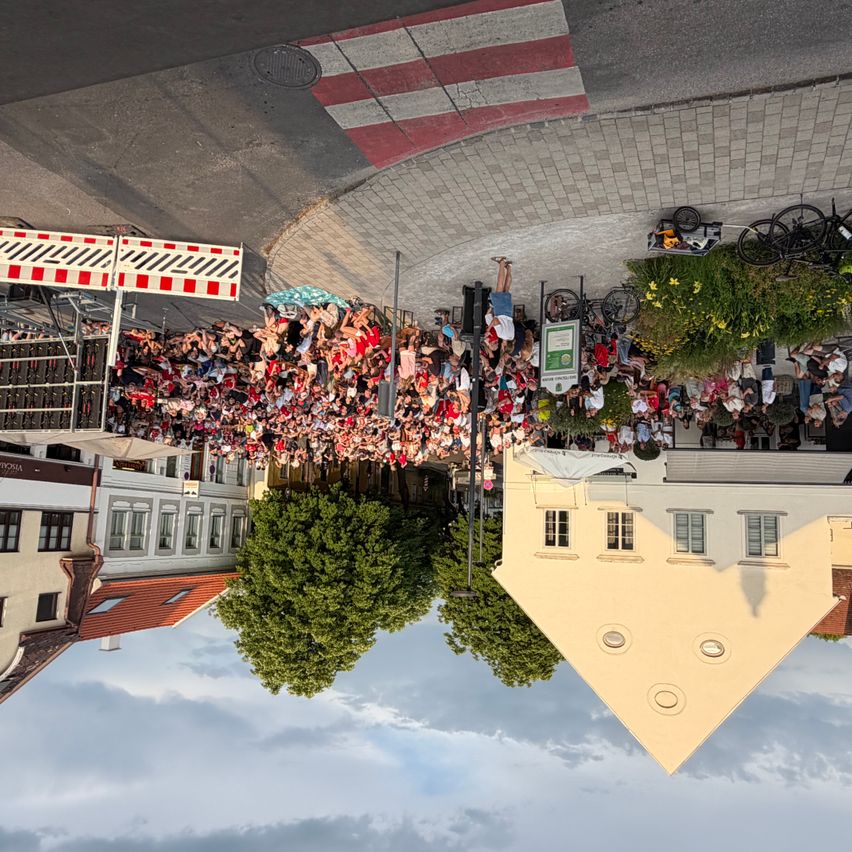 Eine Menschenmenge versammelt sich um ein Gebäude, mit geparkten Fahrrädern in der Nähe. Bäume und Straßenlampen sind sichtbar, und die Szene wird aus einer Vogelperspektive aufgenommen.