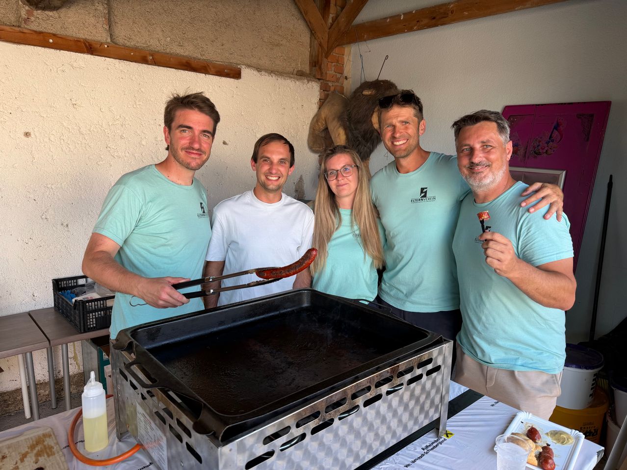 Fünf Menschen in passenden hellblauen T-Shirts posieren für ein Foto. Vor ihnen steht ein großer Grill. Sie halten alle Essen, außer die Frau.