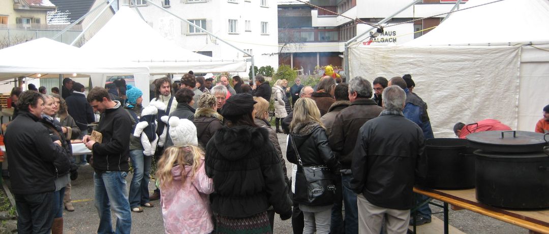 Eine Gruppe von Menschen, darunter Erwachsene und Kinder, versammelt sich um einen Tisch unter einem Zelt auf einem Parkplatz. In der Nähe befinden sich Gebäude, von denen eines ein Schild mit der Aufschrift Haus Balgach trägt.