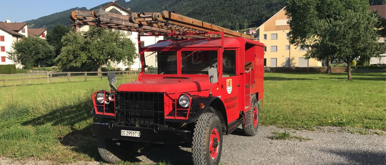 Ein roter Militärlastwagen mit einer Leiter auf dem Dach steht auf einem Kiesplatz mit einem Haus und Bäumen im Hintergrund.