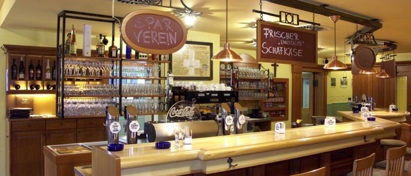 Das Innere einer Bar mit Holzregalen, Flaschen, Gläsern und einer Theke. Es gibt Schilder, die 'SPAR VEREIN' und 'Frischer Ensteller' lesen. Die Theke hat Gläser, Flaschen und eine Coca-Cola-Maschine.