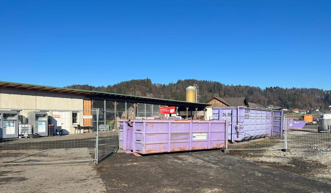 Lila Container stehen in einer Reihe vor einem Gebäude mit grünem Dach, vor dem Hintergrund von Bergen und klarem Himmel.