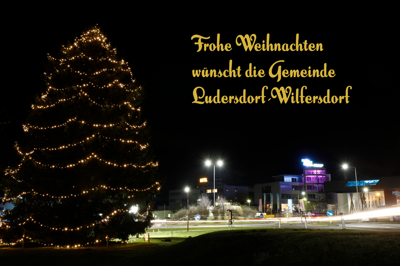 Ein mit Weihnachtslichtern geschmückter Stadtbaum, nachts beleuchtet. Der Textüberlagerung steht 'Frohe Weihnachten', das einen fröhlichen Weihnachtswunsch ausdrückt. Im Hintergrund sind Gebäude mit eingeschalteten Lichtern zu sehen, die eine festliche Atmosphäre schaffen.