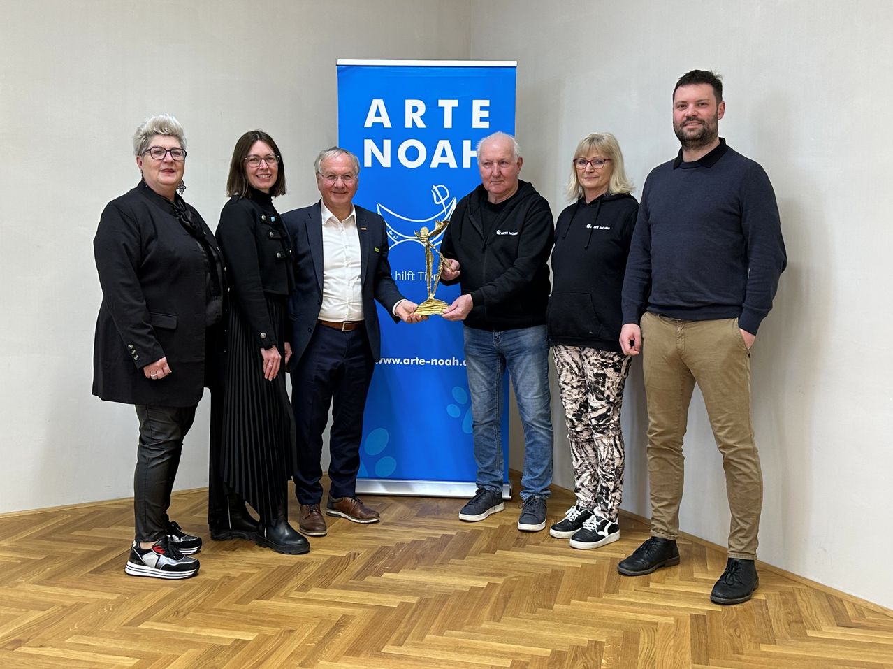 Sechs Personen posieren mit einer Trophäe für ein Foto in einem Raum mit einem blauen Arte Noah-Banner.