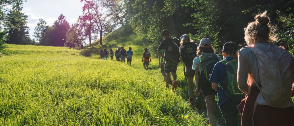 Eine Gruppe von Kindern wandert in einem Grasfeld mit Bäumen, trägt Rucksäcke und Hüte. Die Sonne scheint hell.