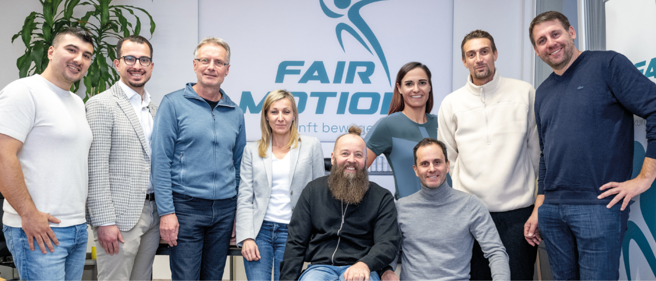 Fünf Personen stehen vor einer weißen Wand mit dem FAIR MOTION Logo. Zwei Frauen und drei Männer, alle lächelnd, scheinen für ein Foto zu posieren.