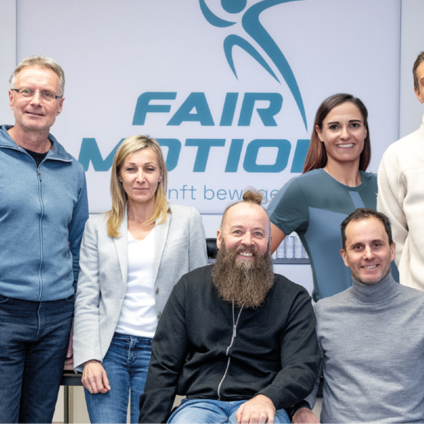 Fünf Personen stehen vor einer weißen Wand mit dem FAIR MOTION Logo. Zwei Frauen und drei Männer, alle lächelnd, scheinen für ein Foto zu posieren.