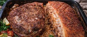 Two roasted pork pieces with herbs and spices on a tray, garnished with rosemary and parsley, accompanied by a bowl of sauce and seasonings.