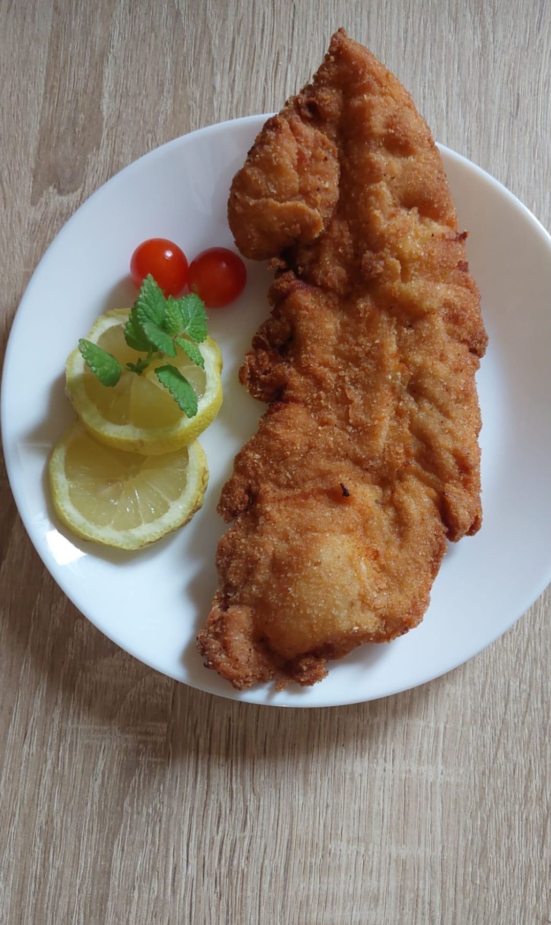 Auf einem weißen Teller befindet sich ein gebratener Fisch mit goldbrauner Kruste, garniert mit Zitronenscheiben und Minzblättern. Zwei Kirschtomaten sind seitlich platziert. Die Umgebung ist eine hölzerne Oberfläche.