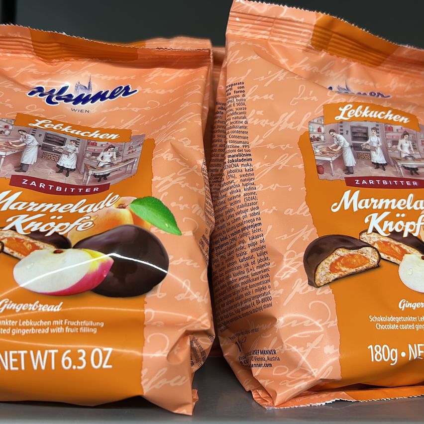 Zwei orangefarbene Tüten Lebkuchen-Gingerbread mit Fruchtfüllung. Eine Tüte ist beschriftet mit 'Marmelade Knotchen' und die andere mit 'Zartbitter Marmelade Knotchen.' Beide haben Abbildungen von Frauen in Schürzen auf der Vorderseite. Gewicht und Nährwertinformationen befinden sich auf der Rückseite.