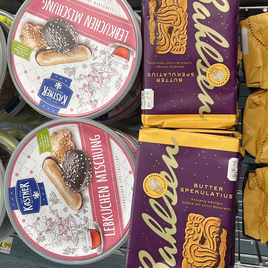 Verschiedene Behälter mit Lebkuchen und Butter Spekulatius sind auf einem Regal ausgestellt. Die Lebkuchen-Behälter haben festliche Designs, während die Butter Spekulatius-Verpackung ein gelbes Etikett mit Text aufweist.