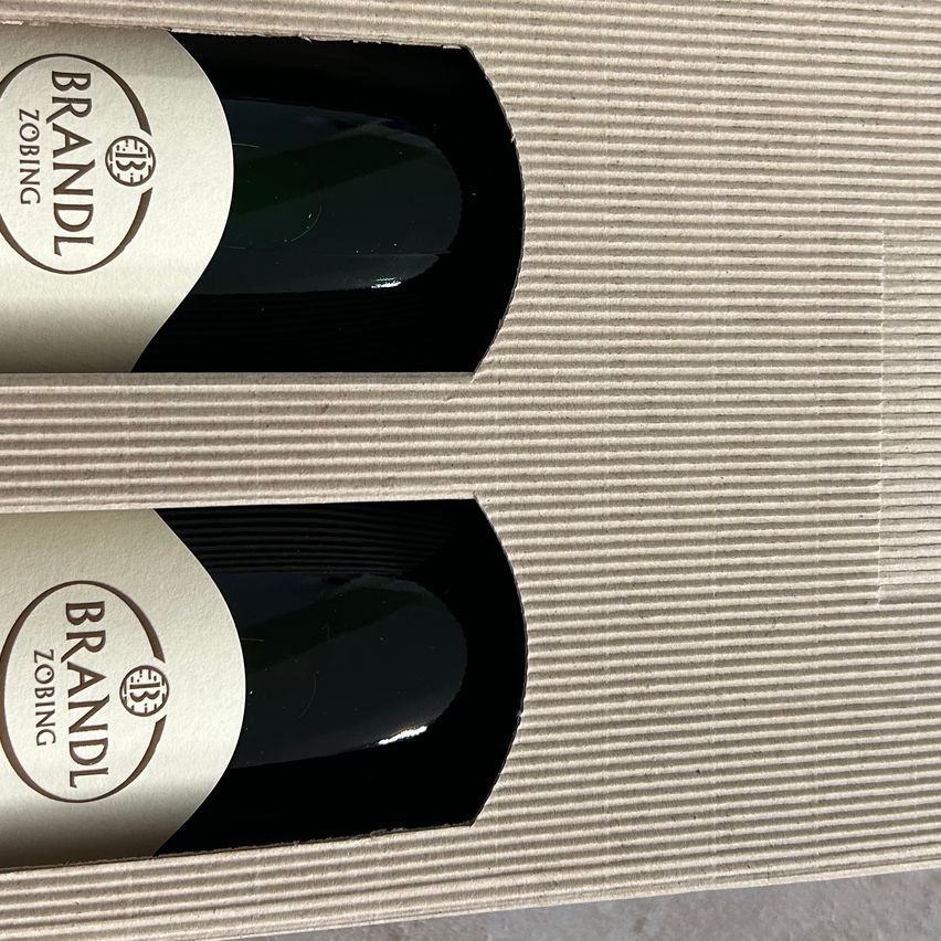 Zwei Weinflaschen in einer Pappschachtel, jede mit einem weißen Etikett, auf dem 'Brandl' und 'Zobing' steht, vertikal positioniert mit einer reflektierenden Oberfläche.