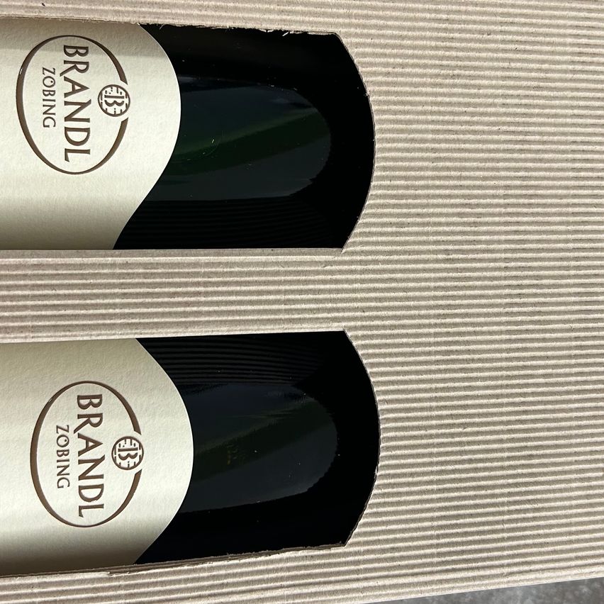 Zwei Weinflaschen in einer Box, beide mit den Etiketten 'Brandl' und 'Zobing', wobei das Etikett ein kreisförmiges Logo und den Text 'BB' in der Mitte aufweist. Die Weinflaschen sind dunkel, und der Hintergrund ist hell.