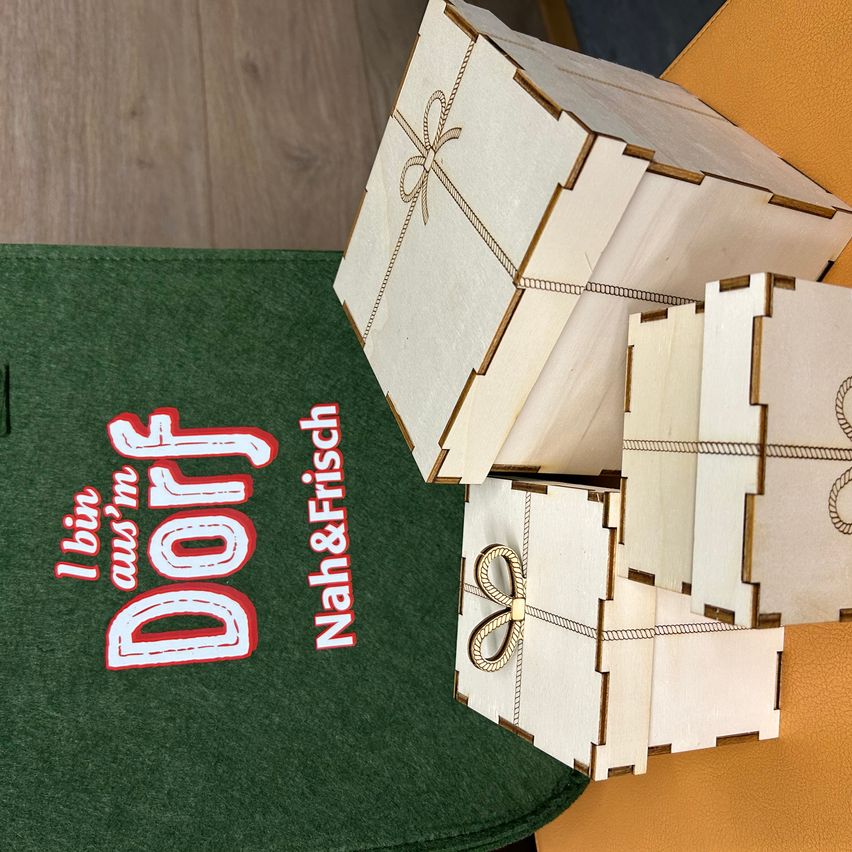 Einige Holzkisten sind auf einem grünen Tuch gestapelt. Die größte Kiste hat ein Band und eine Schleife. Unter den Kisten befindet sich ein grünes Tuch mit weißem Text, der Dorf lautet. Über den Kisten steht das rote Wort Nah&Frisch.