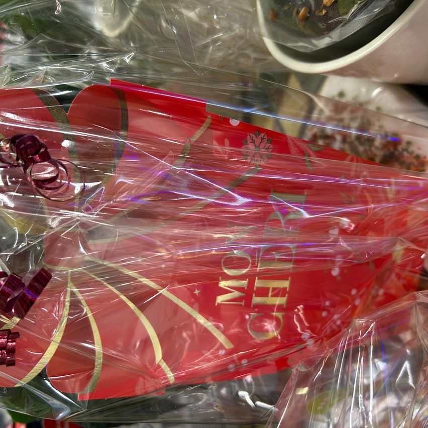 Eine Nahaufnahme von Weihnachtsdekorationen, darunter eine rote Geschenktüte mit 'FROHE WEIHNACHTEN' in Goldfolie geschrieben, in Klarsichtfolie verpackt.