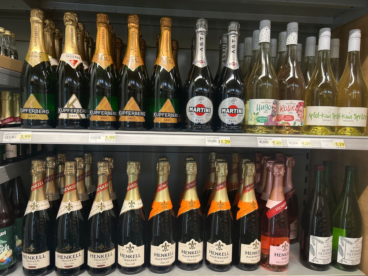 Ein Regal gefüllt mit verschiedenen Schaumweinen, darunter Etiketten wie KUPFERBERG, MARTINI, HUGO und ROSIE, zusammen mit zahlreichen Flaschen von HENKEL TROCKEN. Jede Flasche hat ein eigenes Etikett, und das Preisschild ist auf dem Regal sichtbar.