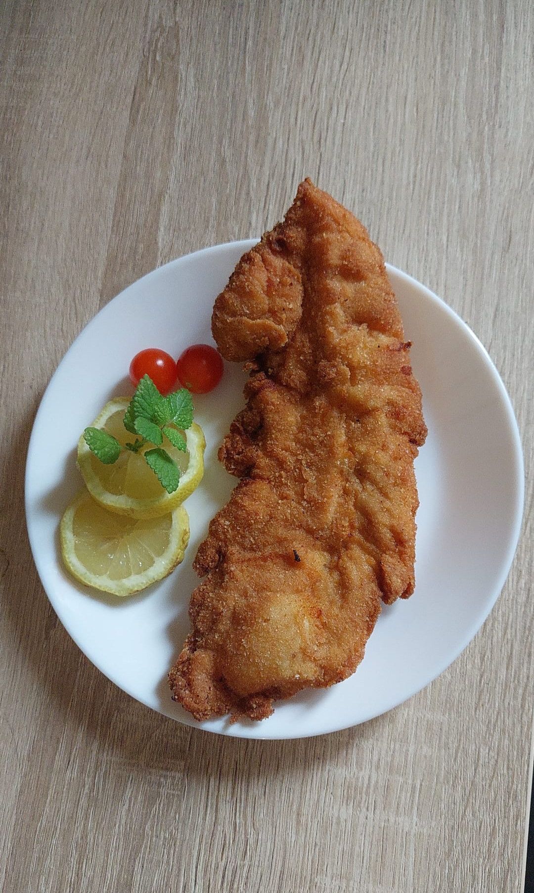 Auf einem weißen Teller befindet sich ein goldbraunes, paniertes Fischfilet, garniert mit Zitronenscheiben und zwei kleinen roten Tomaten. Der Hintergrund ist eine hölzerne Oberfläche.