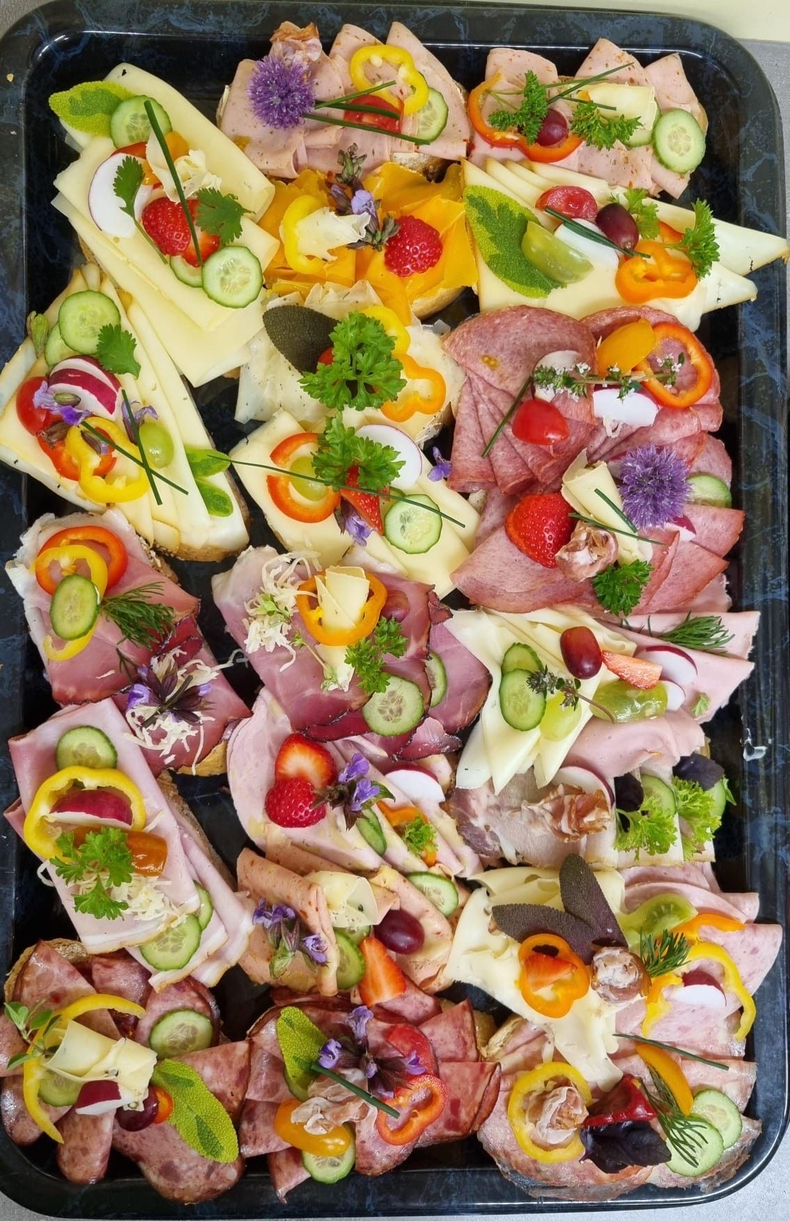 Ein Tablett mit Sandwiches mit Schinken, Käse und Gemüse. Scheiben von Käse, Gurke und Tomate. Jedes Sandwich hat Blumen und Kräuter.