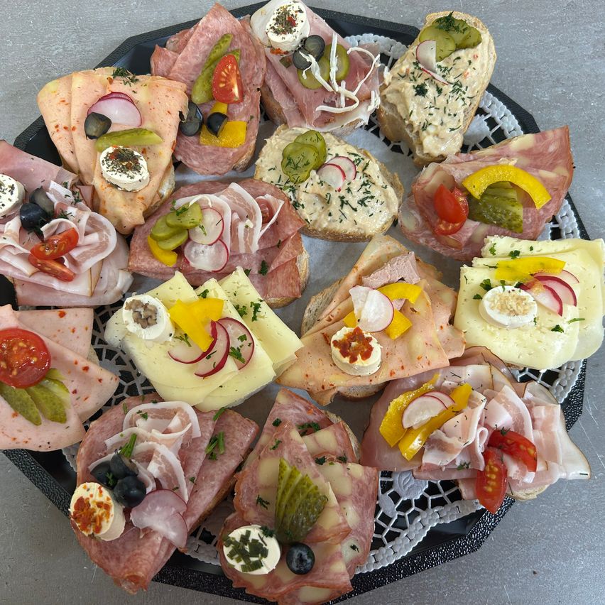 Ein Teller mit verschiedenen Sandwiches, jeweils belegt mit verschiedenen Fleischsorten, Käse und Gemüse. Einige haben eingelegtes Gemüse und Kräuter.