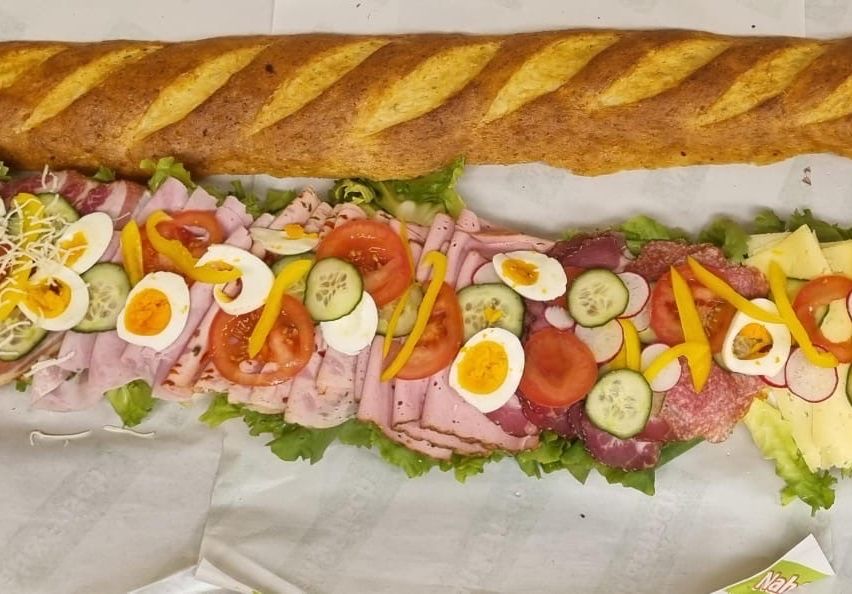 Ein langes Sandwich mit Schinken, Eiern, Gurken, Tomaten und Salat. Obenauf liegt ein Baguette.