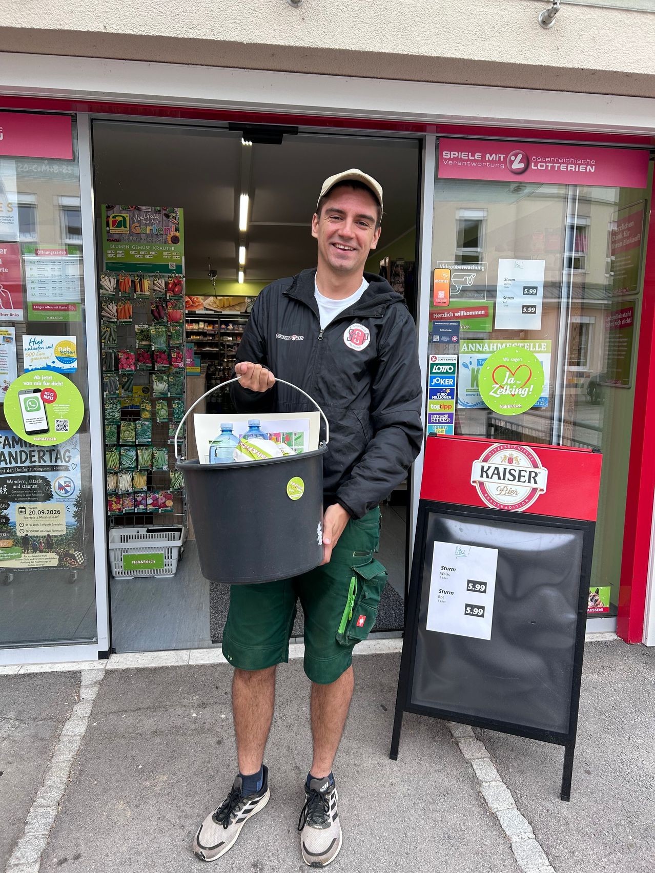 A man in a jacket and green shorts holds a bucket filled with water bottles in front of a store.