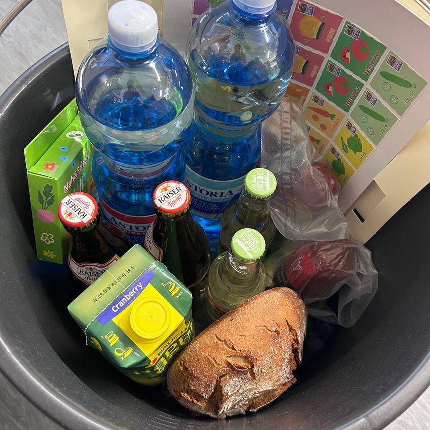 A bucket filled with various items including bottles of water, juice, and wine, a loaf of bread, and a box of flowers.
