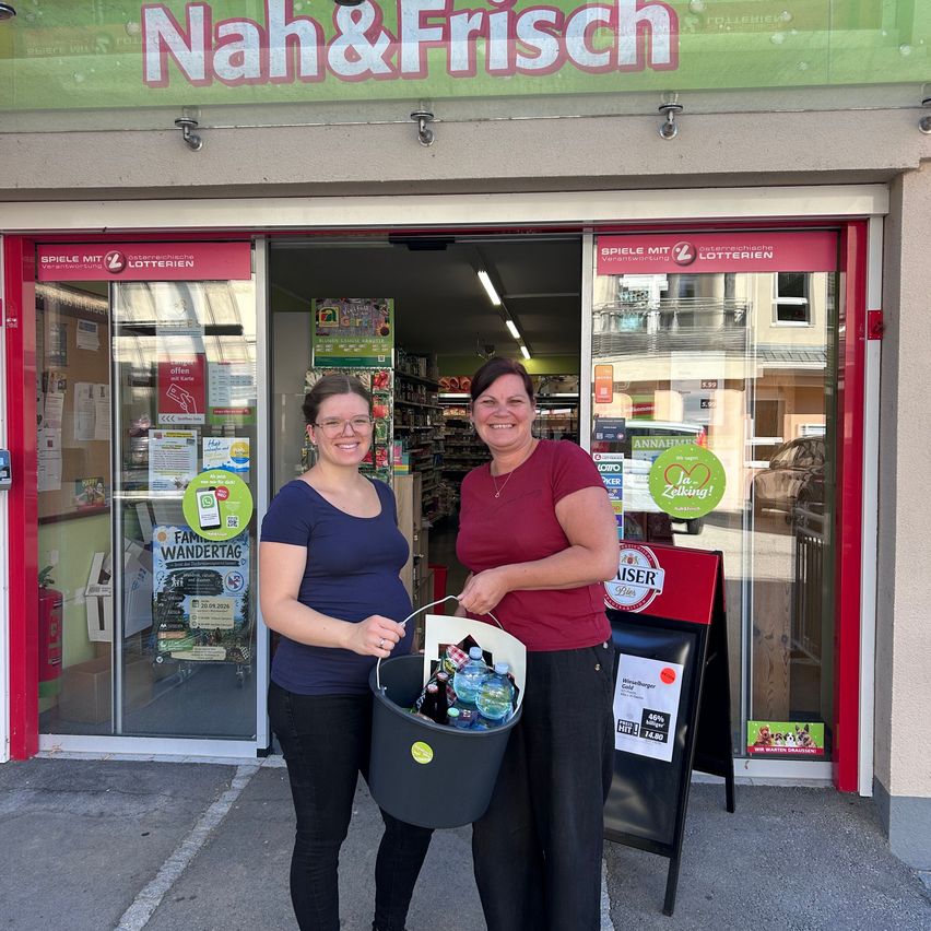 Two women stand outside a store, one holding a bucket filled with water bottles. They are smiling and appear to be posing for a photo. The store has a sign that reads 'Nah&Frisch' and a glass door with a reflection of a building.